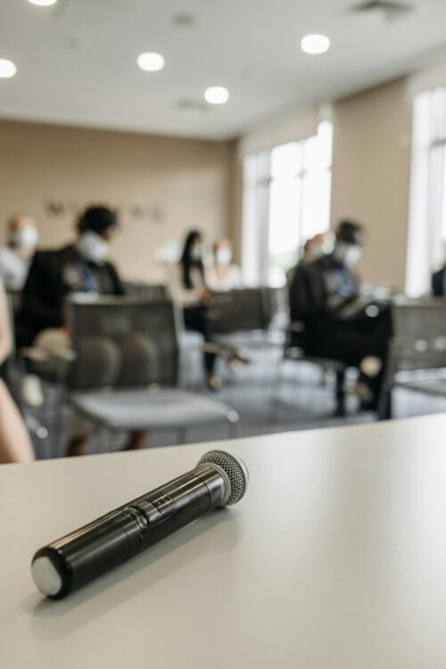 A business conference room setting with masked participants and a microphone in the foreground.